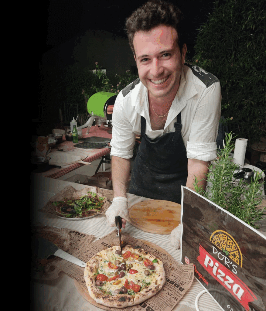 אדם מחייך עומד מאחורי שולחן עם פיצה, סלט ותנור פיצה ירוק, וחותך את הפיצה עם סכין.