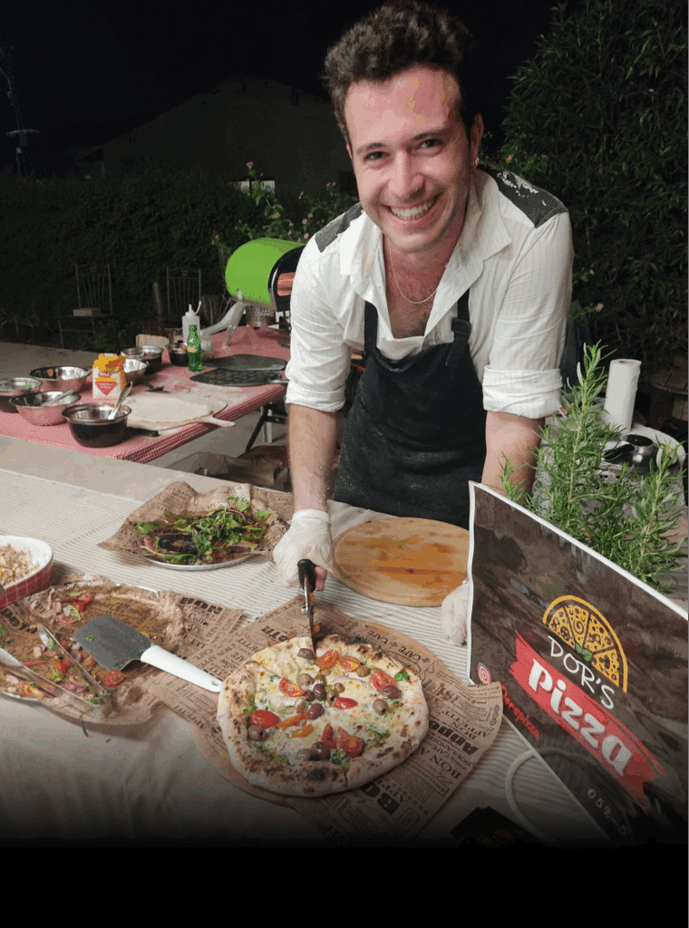 גבר מחייך עומד מאחורי שולחן עם פיצות, סלטים ושלט "פיצה דור'ס".