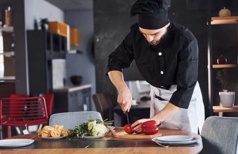 טבח חותך ירקות אדומים על שולחן במטבח, לצד ירקות נוספים ופסטה.