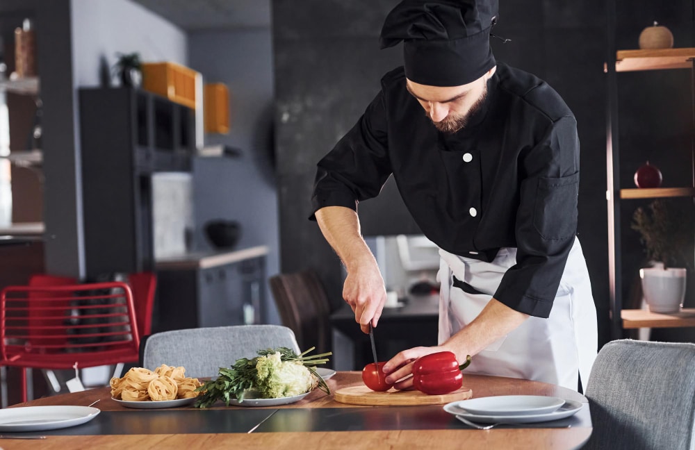 טבח חותך ירקות אדומים על שולחן במטבח, לצד ירקות נוספים ופסטה.