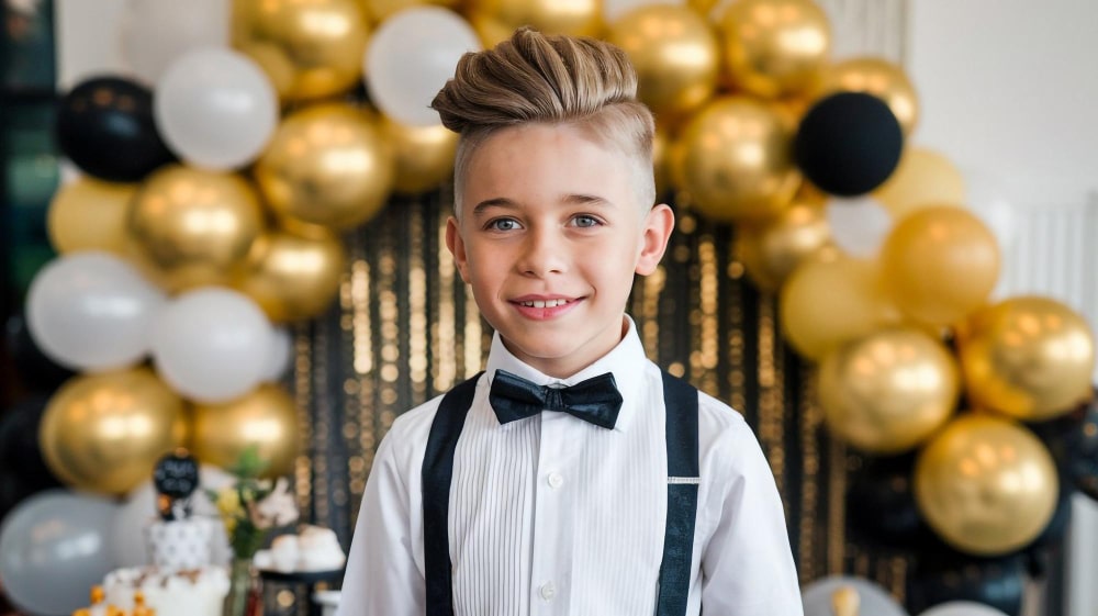 ילד מחייך, לבוש בחולצה לבנה, פפיון וכתפיות שחורות, עומד מול רקע של בלונים בצבעי זהב, שחור ולבן.