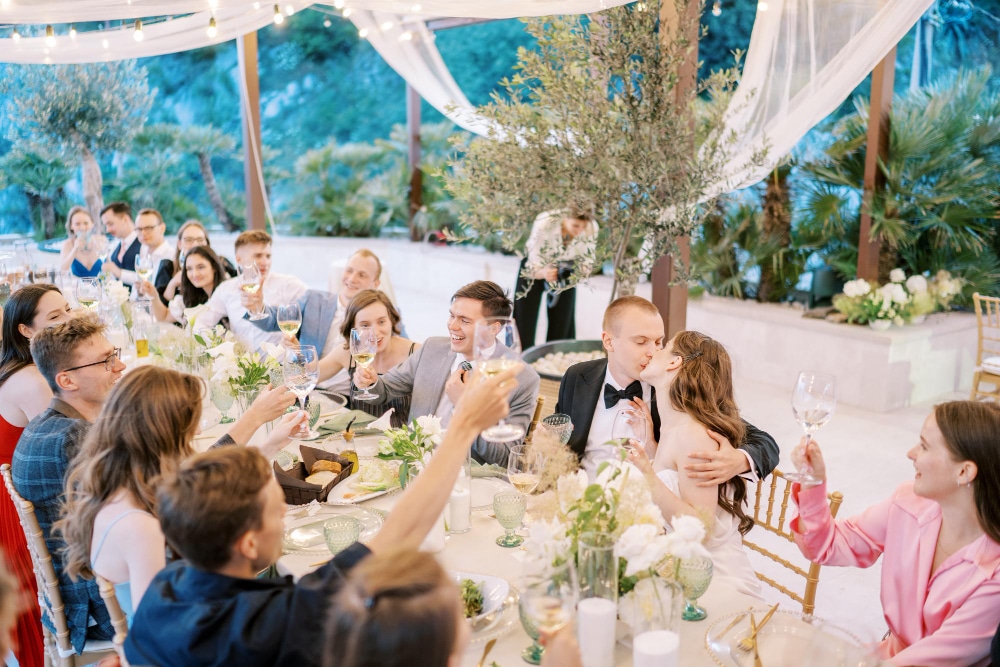 אורחים יושבים סביב שולחן ארוך ומלא כלים, מרימים כוסות יין ומחייכים, בעוד הכלה והחתן מתנשקים.