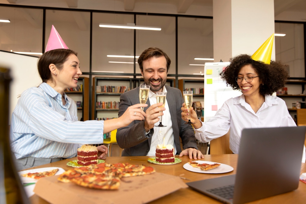 שלושה אנשים חוגגים עם כובעי מסיבה, פיצה, עוגות וכוסות שמפניה מול מחשב נייד.