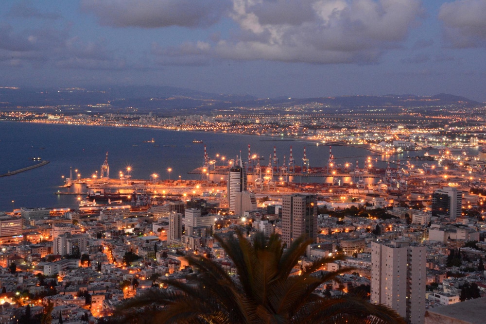 מבט לילי על עיר נמל מוארת, עם מנופים בנמל, בניינים מוארים וים כחול כהה ברקע.