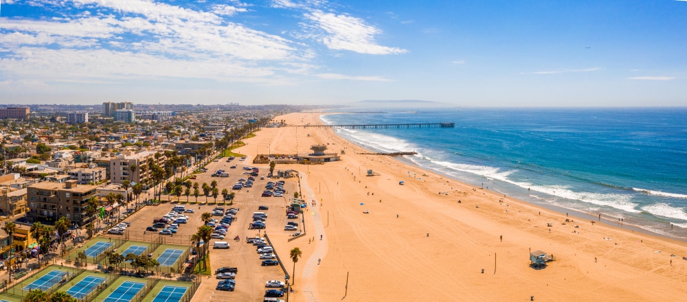 מבט אווירי על חוף ים חולי עם אנשים, מגרשי טניס, חניון מלא מכוניות, עיר ברקע ומזח בים.