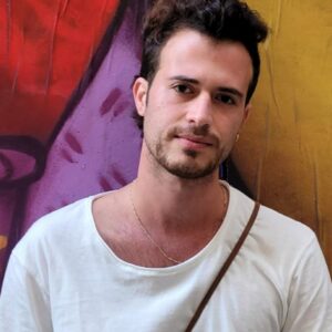 גבר צעיר עם שיער כהה וחולצה לבנה עומד מול קיר צבעוני עם גרפיטי.