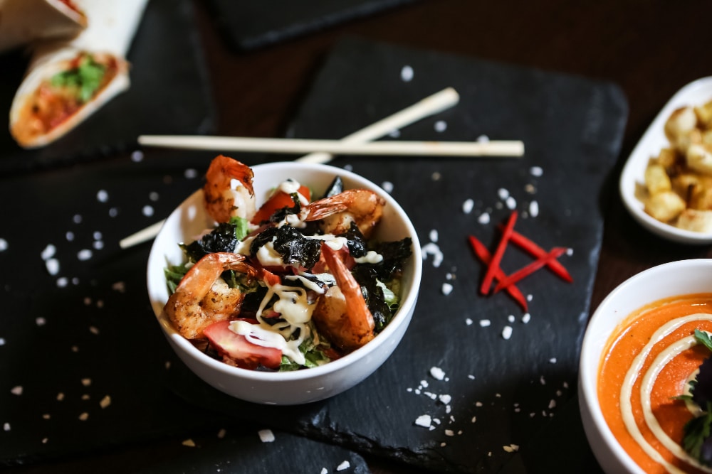 קערת סלט עם שרימפס צלויים, אצות, רוטב וירקות, מוגשת לצד מנה אחרת ומקלטי אכילה.